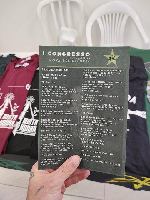 Folder do evento na banca de venda de merchandising, incluindo a camiseta da Nova Resistência com o lema de Mussolini